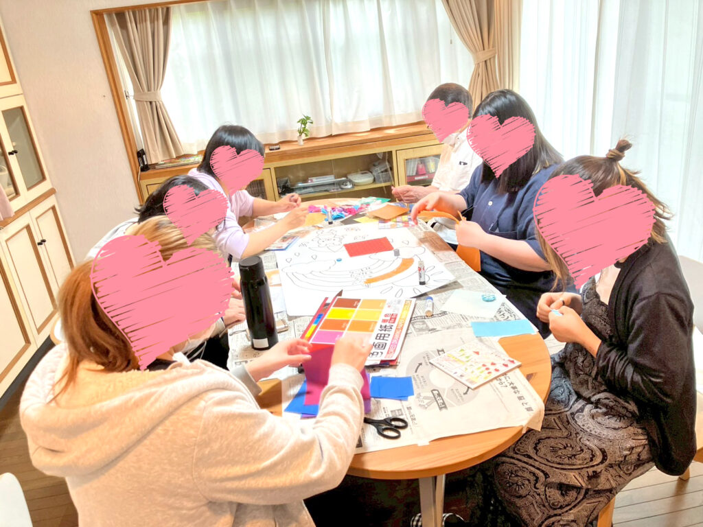 6人の入居者とスタッフがテーブルを囲み、折り紙を使ったちぎり絵の壁飾り作りに取り組んでいます。新聞紙を敷いたテーブルには色とりどりの紙や道具が並び、和やかな雰囲気です。