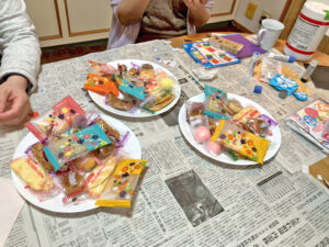 新聞紙を敷いたテーブルの上に、お茶会用の個包装のお菓子が盛られたお皿が3枚並んでいます。せんべいや洋菓子などがあり、賑やかで楽しそうな雰囲気です。