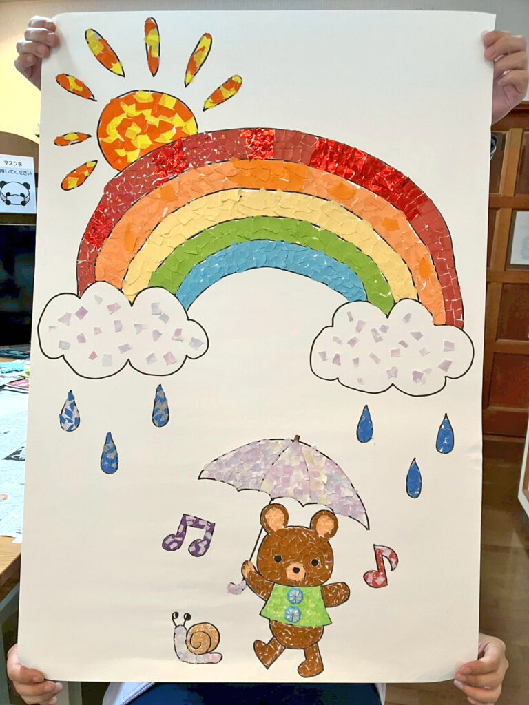 仕上がった壁飾りを持っている様子です。傘をさしたくまや虹、雨粒、音符などが色とりどりに貼られており、明るく楽しい作品に仕上がっています。