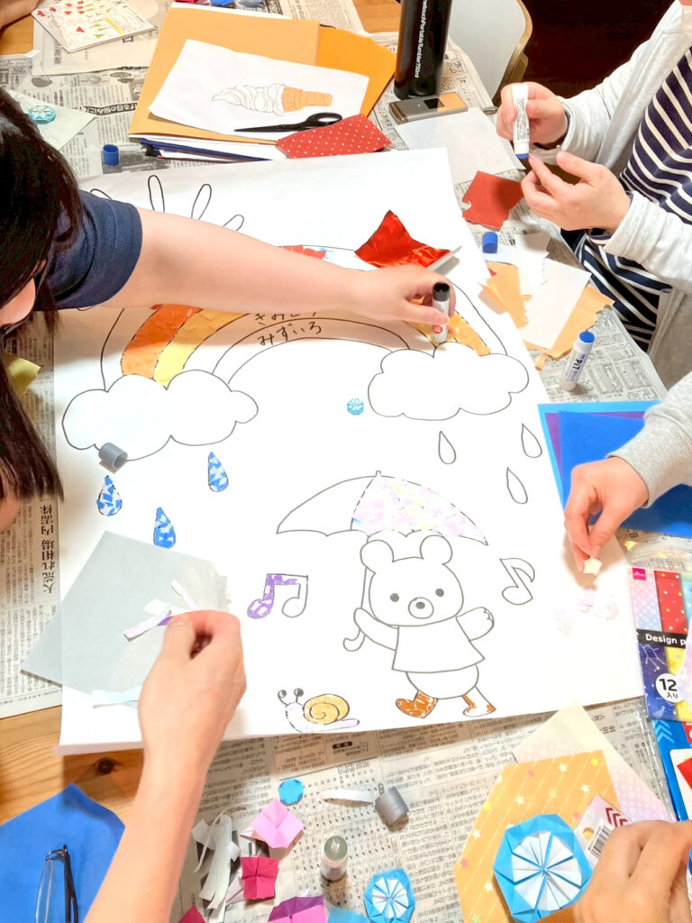 模造紙に描かれた下絵に、折り紙をちぎって貼っている手元の様子です。傘をさしたくまや、雨粒や音符、虹なども見え、賑やかな雰囲気の壁飾りが出来上がってきています。