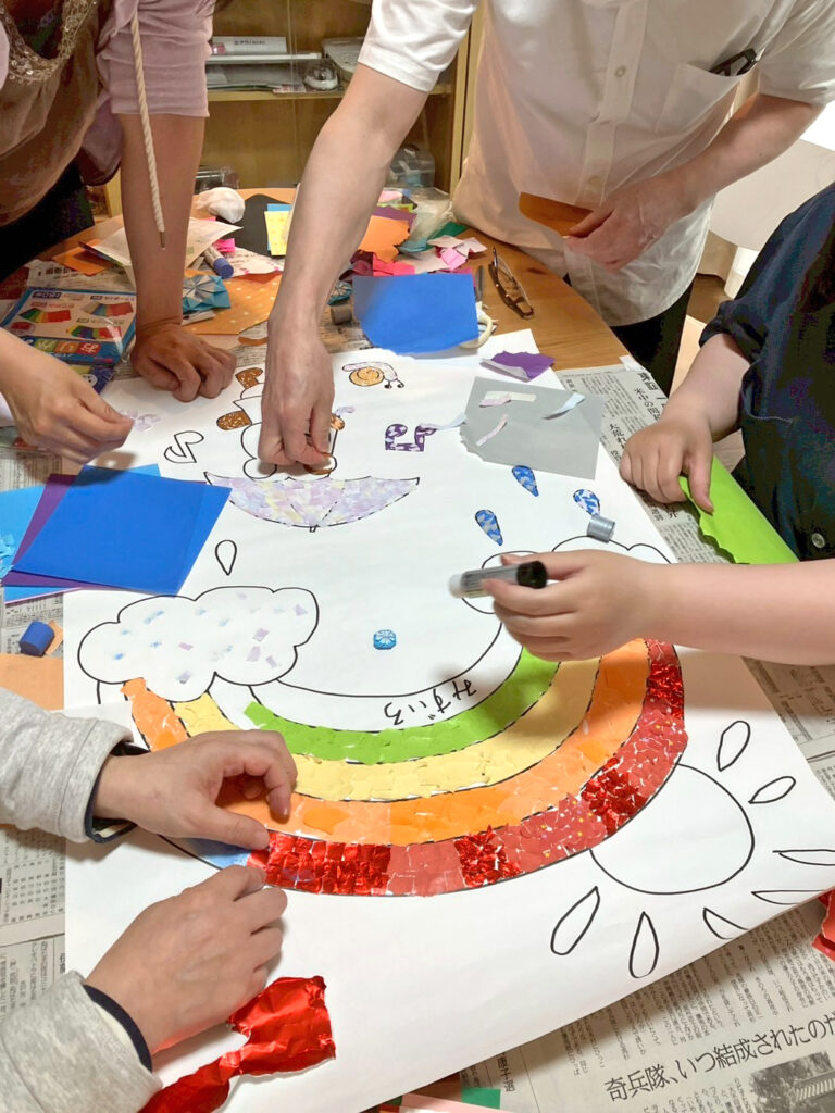 色とりどりの折り紙で作られたちぎり絵を、数人の手で仕上げている様子です。赤や黄色の折り紙が貼られ、少しずつ完成に近づいている場面です。