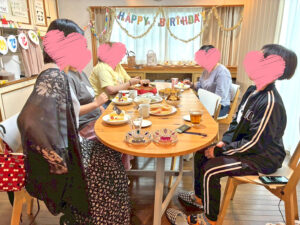 丸テーブルを囲んで5人が歓談している様子。テーブルにはケーキやお菓子が並び、奥の壁には「HAPPY BIRTHDAY」のガーランドが飾られている。